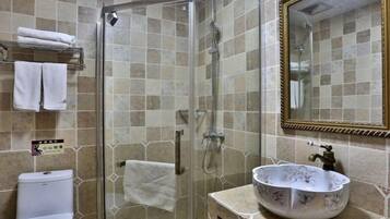 Un baño con ducha de vidrio, inodoro blanco, lavamanos con un recipiente decorativo y un espejo con marco dorado.