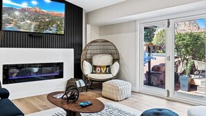 Una sala de estar moderna con un televisor de pantalla plana, una chimenea, un sillón cómodo con un cojín que dice 'LOVE' y un amplio ventanal corredizo que da a un espacio exterior.
