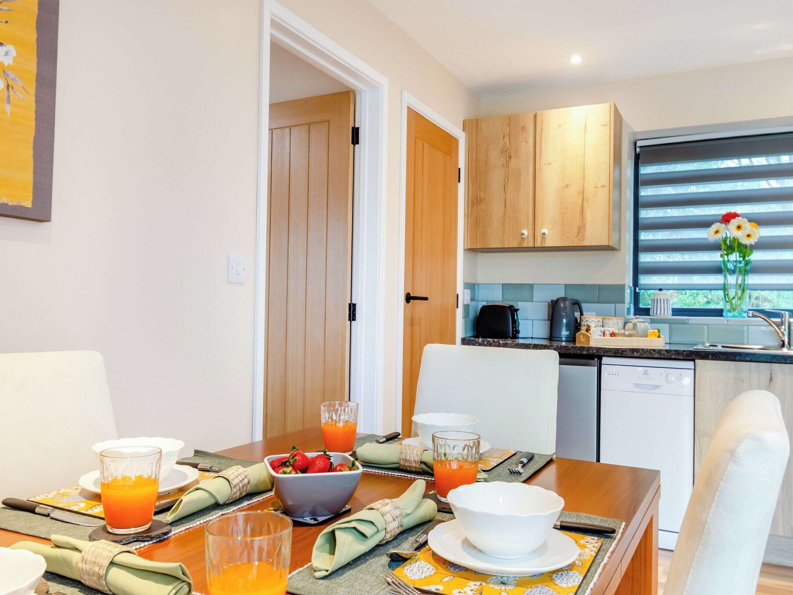 A modern kitchen with wooden cabinets, a dining table set for breakfast with fruit and juice, and a window with blinds.