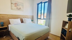 A bedroom with a large bed, a wooden headboard, a painting on the wall, a window with curtains, a TV on the wall, and a small plant on a shelf. - Sea Glass Sunset (Rafailovići)