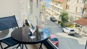A balcony with a round table and two chairs, overlooking a street with parked cars and buildings. - Sea Glass Sunset (Rafailovići)