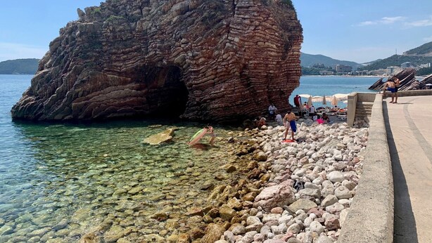 A rocky coastline with clear water, a large rock formation, and a paved walkway with people. - Sea Glass Sunset (Rafailovići)
