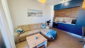 A modern living room with a beige sofa, a wooden coffee table, and a kitchen area with blue cabinets and a wooden countertop. - Sea Glass Sunset (Rafailovići)
