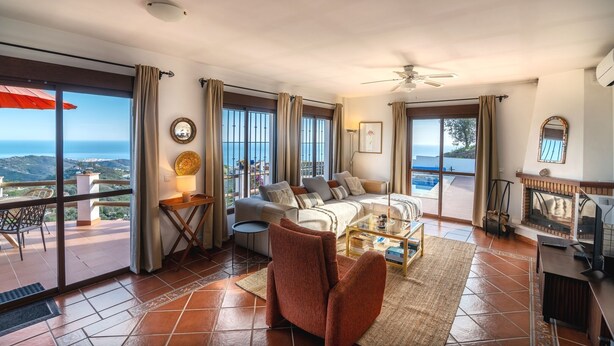 A spacious living room with a sofa, armchairs, and a coffee table. There is a fireplace, a ceiling fan, and large windows offering a view of the sea. - Los Naranjos - Luxury villa with private pool and stunning sea view (Sayalonga)