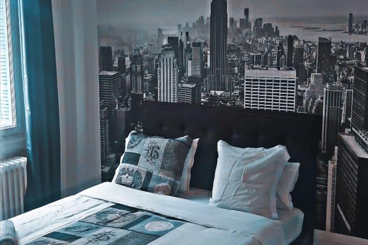 A bedroom with a large bed, a cityscape mural on the wall, and a window with curtains.