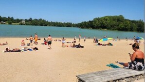 Una playa con gente tomando el sol y nadando, rodeada de árboles y un lago tranquilo.