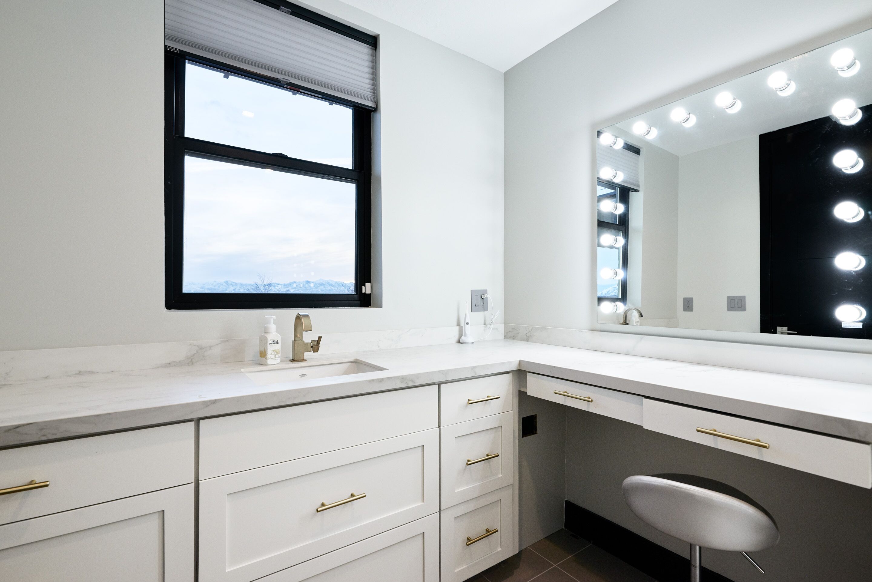 A modern bathroom with a large vanity, a mirror with lights, a window, and a chair.