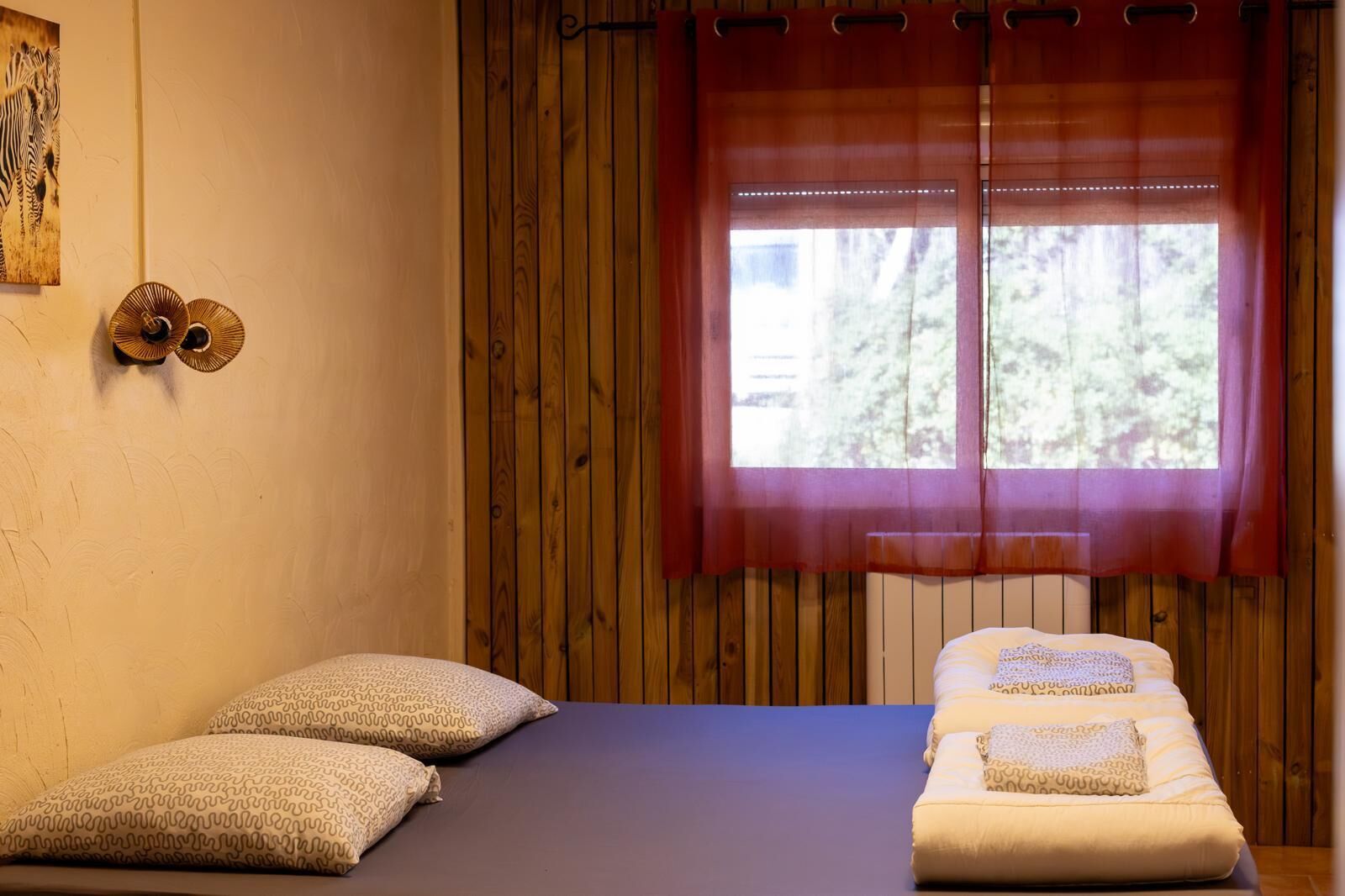 Una habitación con una cama, dos almohadas, un ventanal con cortina roja y una pared de madera.