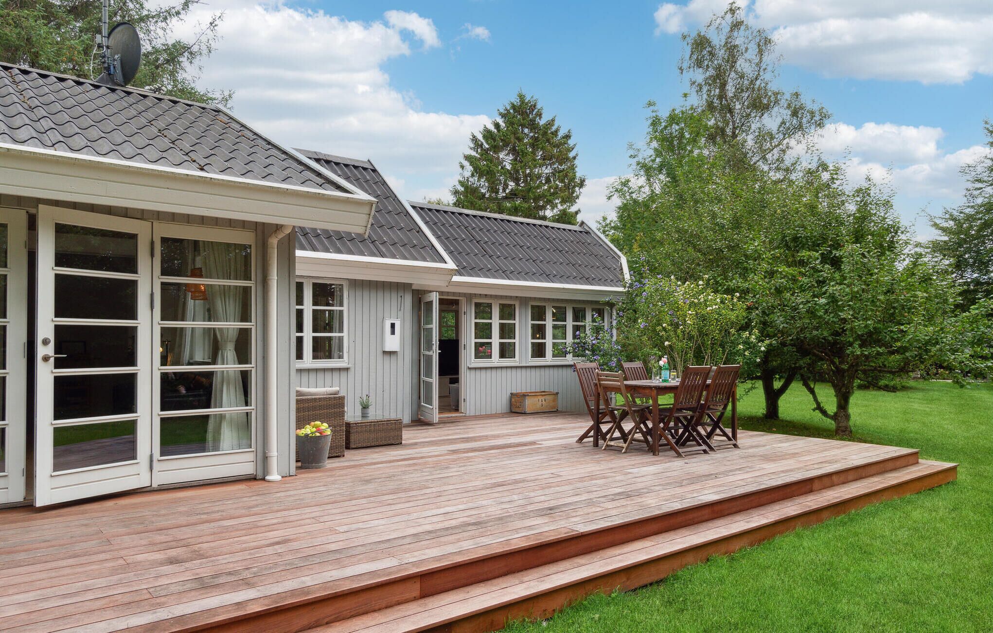 Een huis met een terras, een eethoek met stoelen en een tafel, en een groot raam met uitzicht op een gazon.