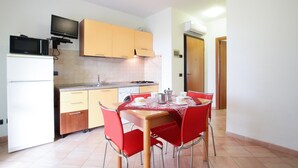 Een praktische keuken met een magnetron, kookplaat en spoelbak. Er staat een eettafel met rode stoelen en een tafelkleed.