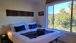 A neatly made bed with white and dark blue bedding, a bedside lamp, and a window with a view of trees and hills. - Magnificent Sedona Redrock Views; 7-Min Walk to Uptown;Single-Level;Eco-Friendly (Sedona)