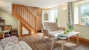 A cozy living room with a wooden staircase, a patterned sofa, a coffee table with books and a vase, and a window with a view of greenery. - 3 bedroom accommodation in St Austell (St Austell)