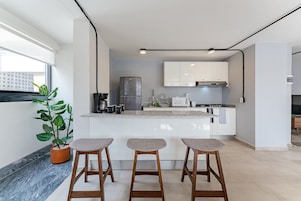 Une cuisine moderne avec des armoires blanches, un réfrigérateur en acier inoxydable et un four encastré. On y trouve trois tabourets en bois et une plante en pot.