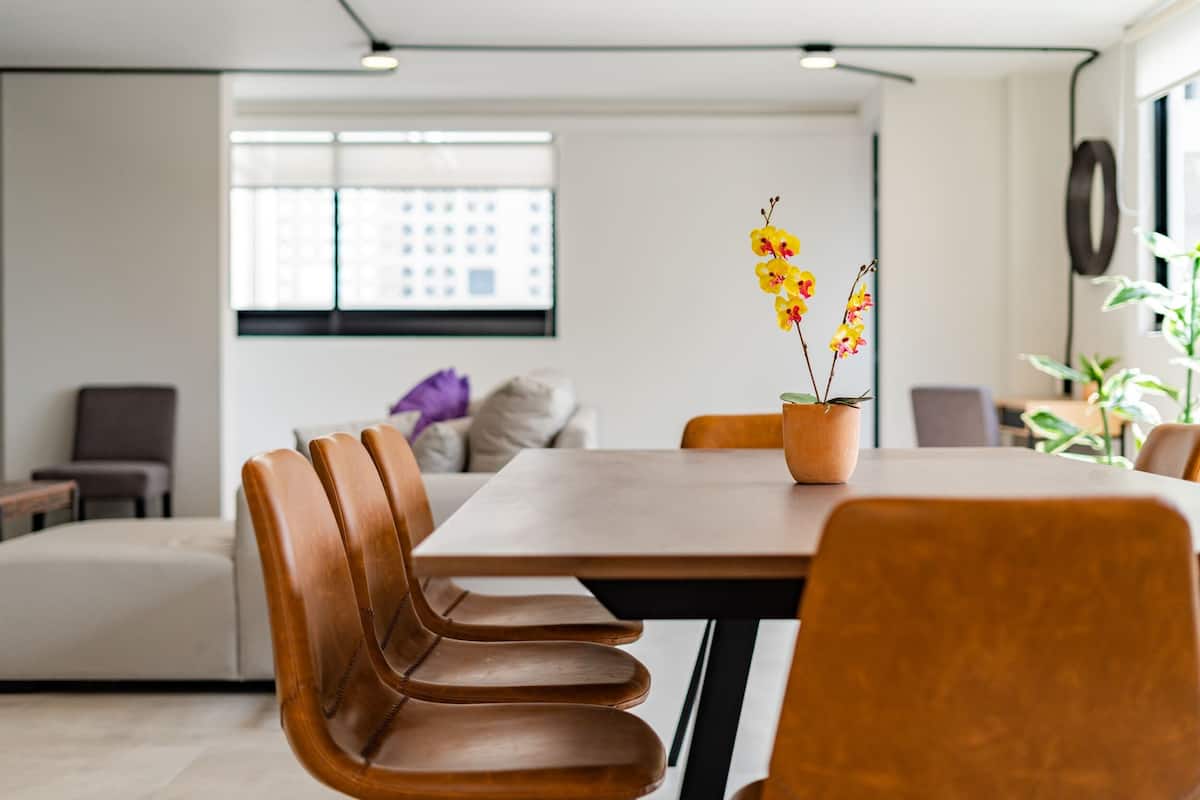 Un comedor moderno con una mesa y sillas de madera, una orquídea en maceta y vistas a la ciudad a través de amplias ventanas.