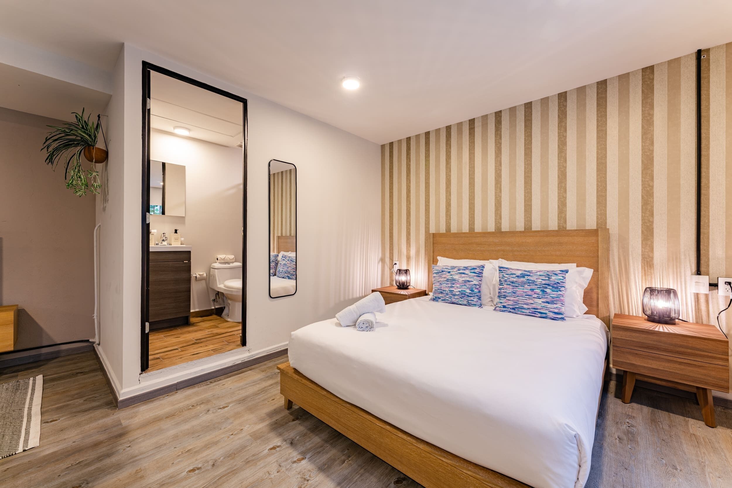 A modern bedroom with a large bed, wooden headboard, bedside tables, a mirror, and striped wallpaper.