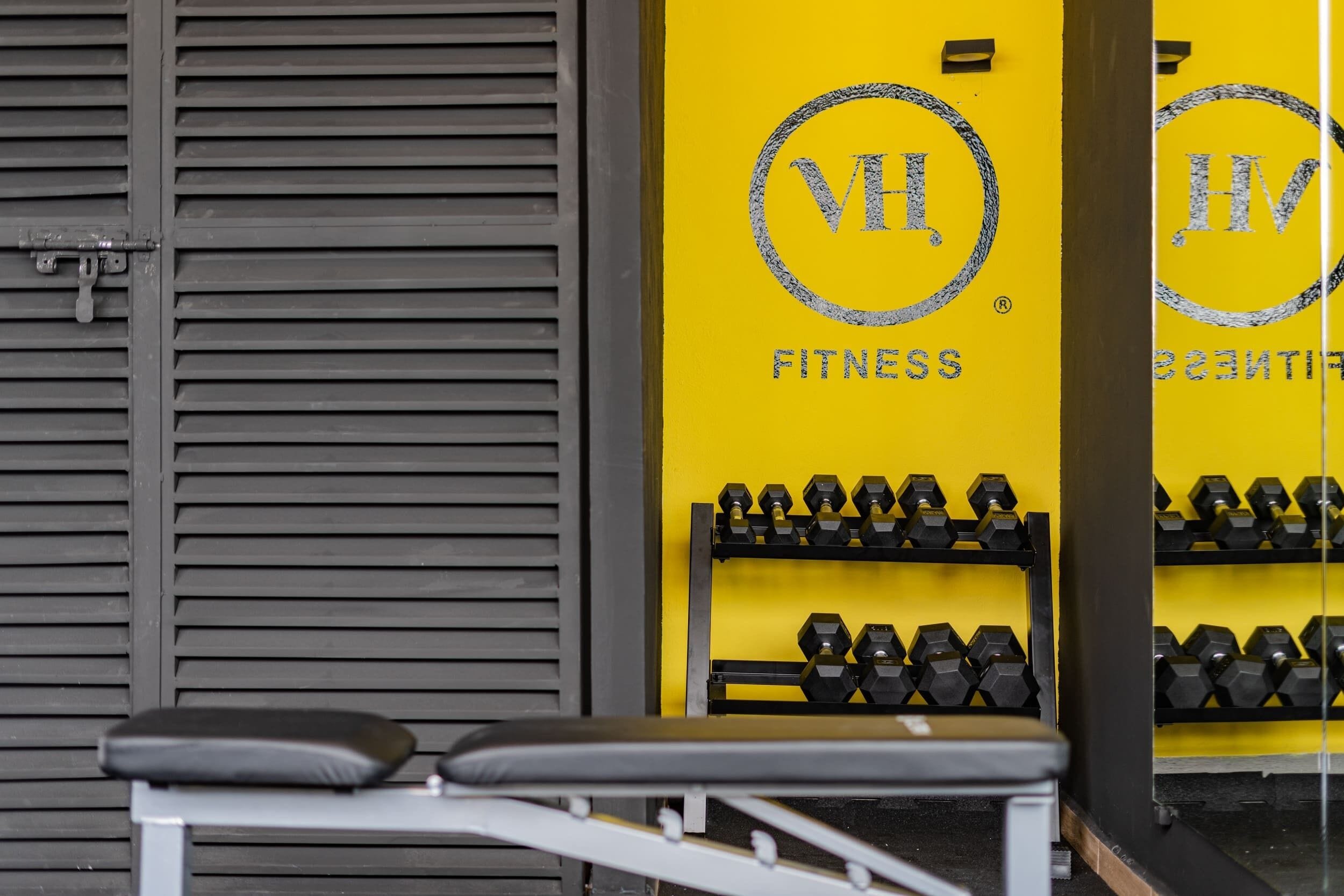 A gym with a bench, dumbbells, and a yellow wall with the logo "VHL FITNESS".