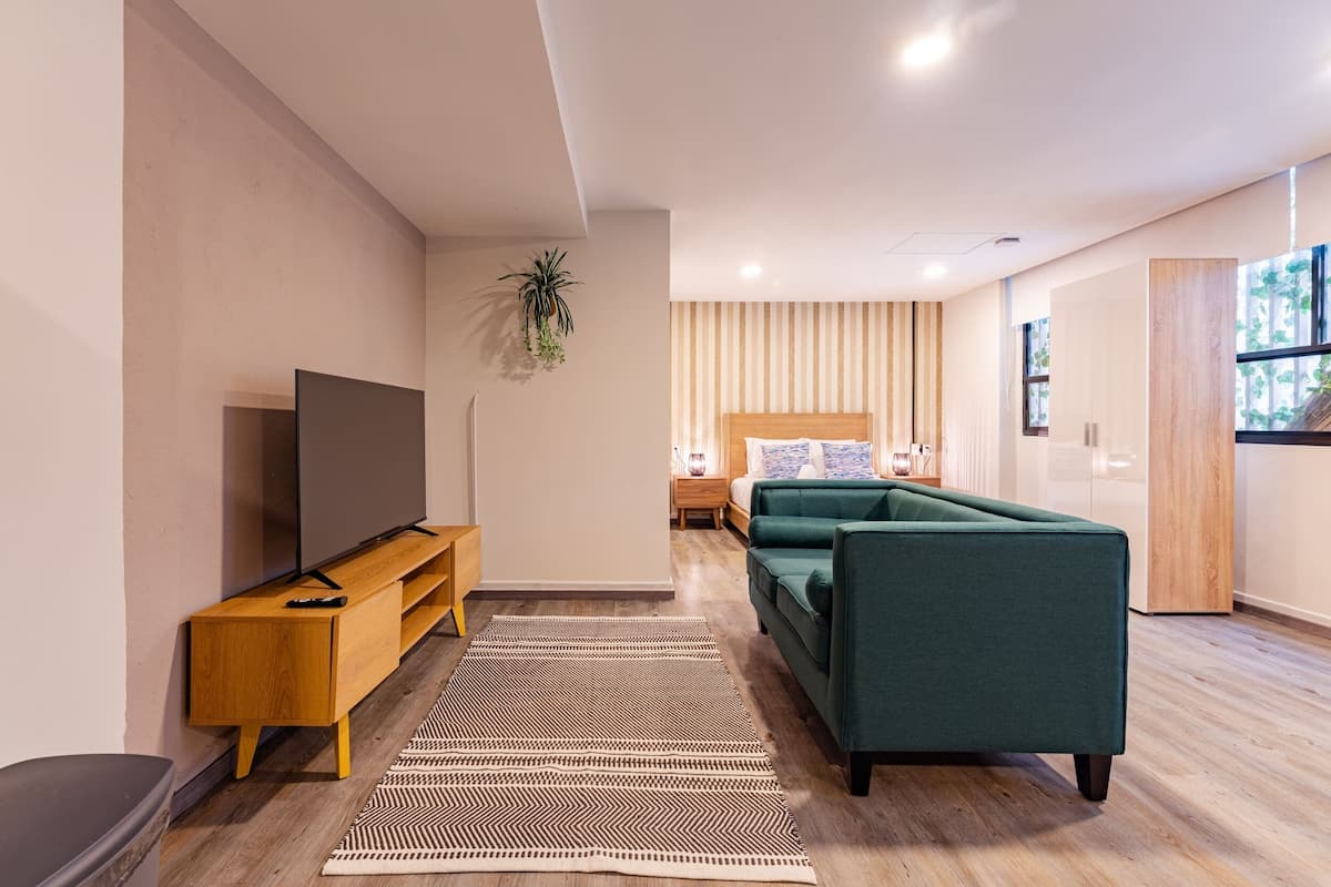 Una sala de estar moderna con un televisor de pantalla plana sobre una consola de madera, un sofá verde, un tapete a rayas y una cama con cortinas a rayas al fondo.