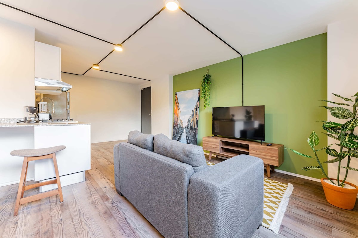 Un espacio habitable moderno con un sofá gris, un mueble de televisión de madera, una zona de cocina y una pared de acento verde.