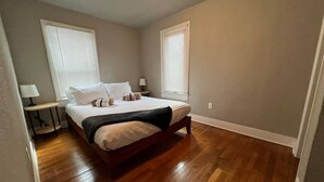 A bedroom with a wooden floor, a bed with white bedding, two bedside tables, and a window with blinds.