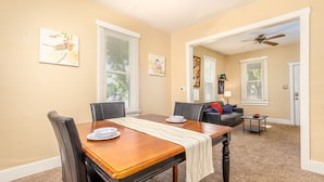 A dining area with a wooden table, black chairs, and a beige carpet. There are two windows with blinds, a ceiling fan, and a sofa in the background.