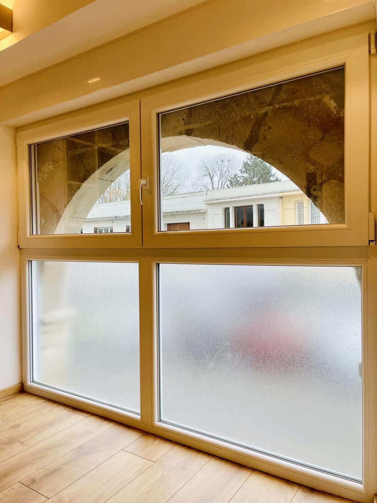 A room with a large window featuring a frosted lower section and a clear upper section, wooden flooring, and a visible building outside.