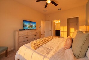 Un dormitorio con cama, televisor sobre un aparador, ventilador de techo y puerta que da a otra habitación.