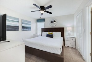 A bedroom with a bed, a ceiling fan, a bedside table with a lamp, and artwork on the walls.
