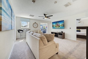 A living room with a beige sofa, a TV, a ceiling fan, and a clock on the wall. - Outstanding 8BR Resort Pool Home Near Disney World (Four Corners)