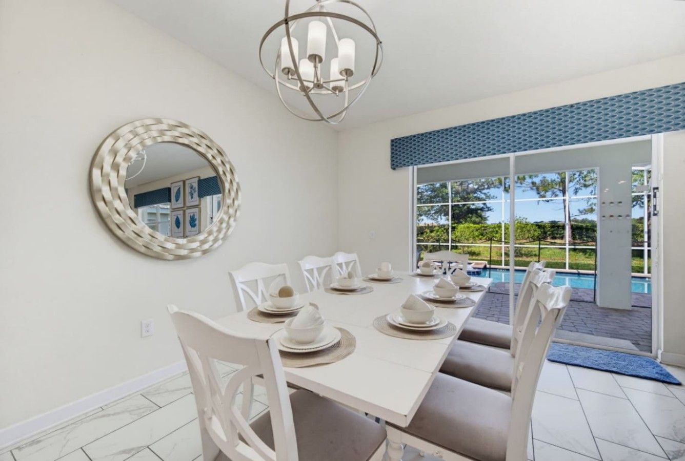 Un comedor con una mesa blanca dispuesta para seis personas, sillas blancas, un espejo redondo y vistas a una piscina a través de grandes puertas de vidrio.