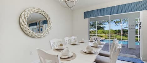 Un coin repas avec une table blanche dressée pour six personnes, des chaises blanches, un miroir rond et une vue sur une piscine, visible à travers de grandes portes vitrées.