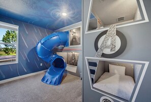 A room with a blue slide, a bed with a headboard, and a window with a view of greenery.