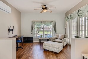 Una habitación con ventilador de techo, sofá, reposapiés, televisor y ventana con cortinas.