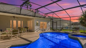 A swimming pool with a curved design, surrounded by a covered patio area with chairs and a table. - The Marvel Mansion 5BR Pool Home Near Disney World (Four Corners)