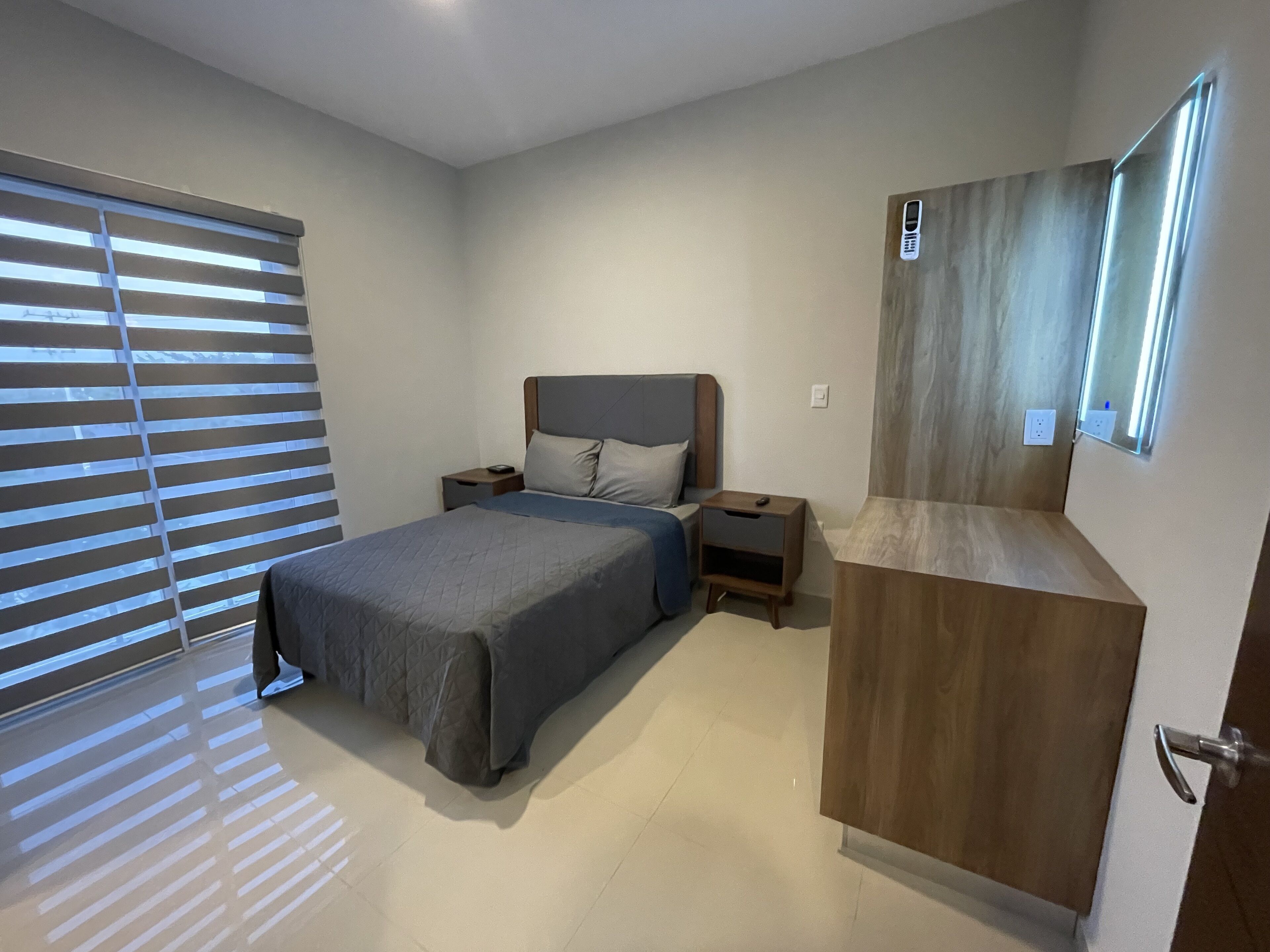 Un dormitorio con cama, mesitas de noche, un armario de madera y una ventana con persianas.