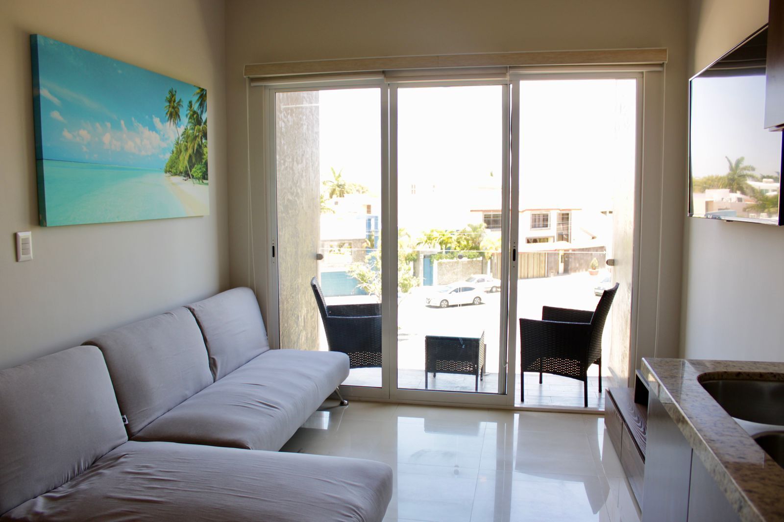A modern living room with a sofa, a dining area with chairs, a kitchen with a sink, and a sliding glass door leading to an outdoor area with a view of buildings and parked cars.