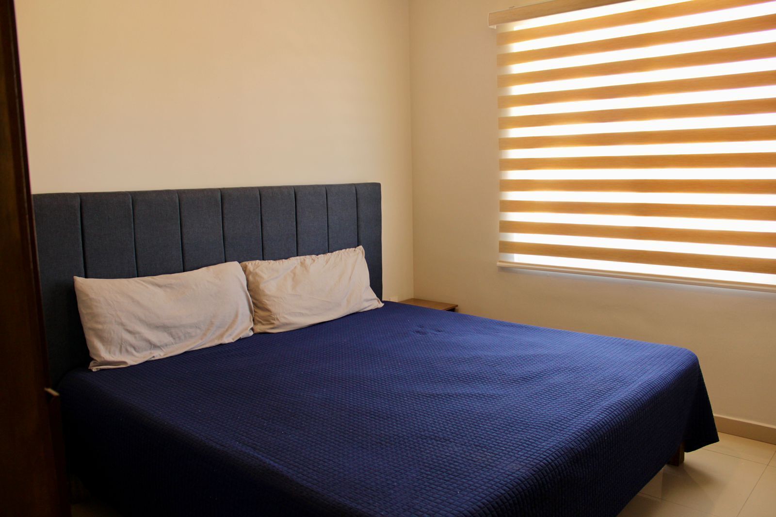 A bed with a dark headboard and two pillows, a window with horizontal blinds, and a plain wall.