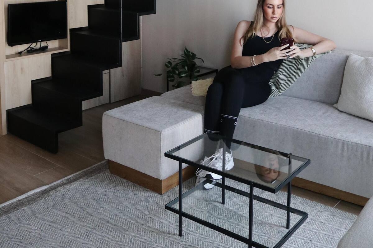 Una sala de estar moderna con un sofá, una mesa de centro de vidrio, un televisor, una escalera y una obra de arte enmarcada en la pared.