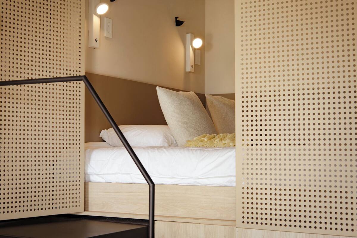 Una cama con ropa blanca, cabecera de madera y dos lámparas fijadas a la pared.