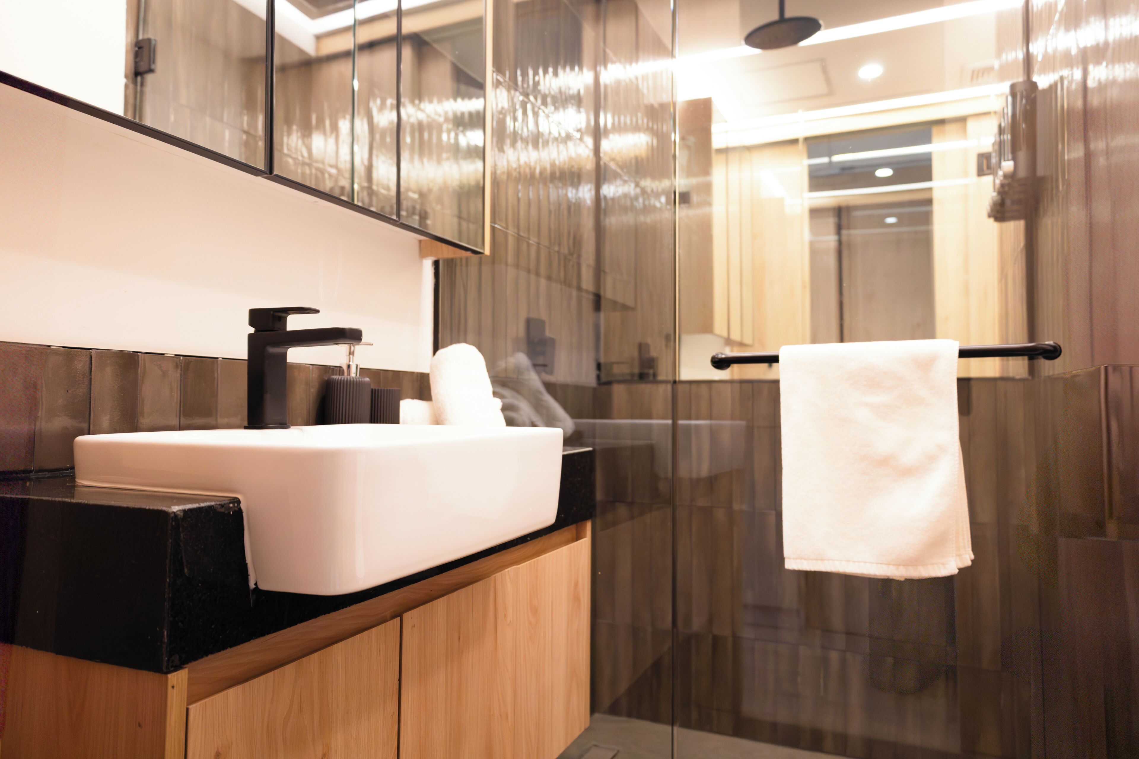 Een moderne badkamer met een witte wasbak, een zwart aanrecht, een houten kast, een glazen douchecabine en een handdoek aan een zwarte handdoekhouder.