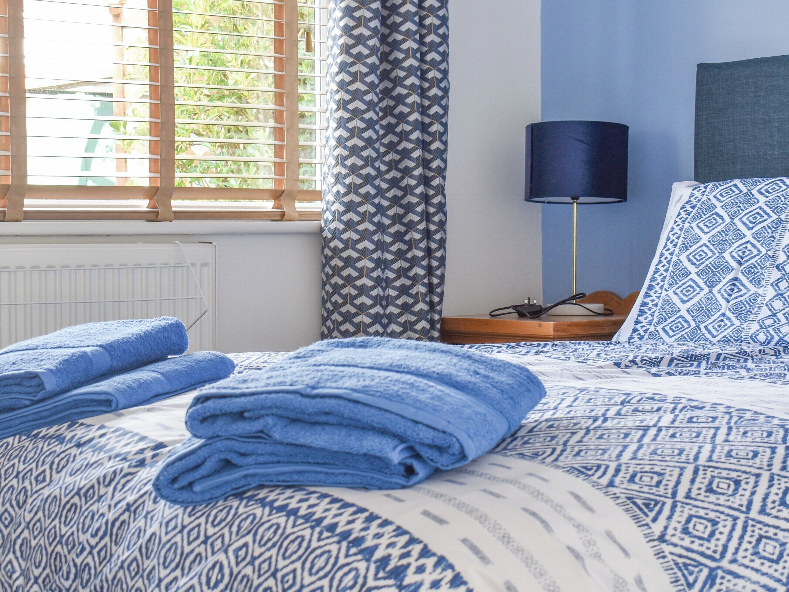 Una cama bien hecha con toallas azules, una colcha azul y blanca estampada, una lámpara de noche y vistas a la vegetación a través de persianas.