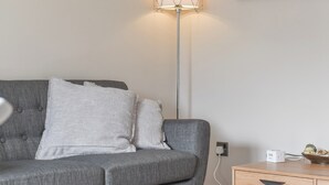 Un canapé gris avec deux coussins, une lampe sur pied, une table d’appoint en bois sur laquelle se trouve un appareil blanc, et une prise électrique murale.