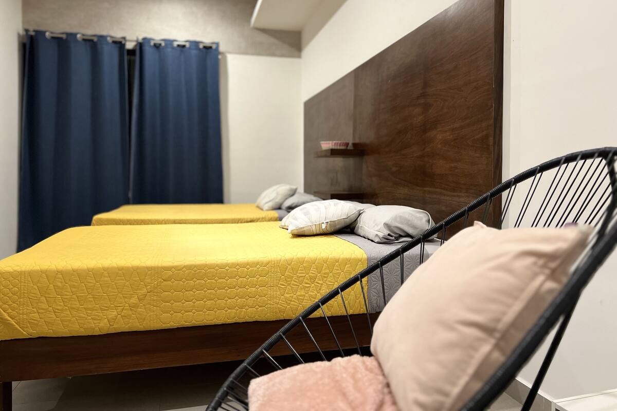 Una habitación con dos camas, un cabecero de madera, una silla negra y una cortina azul.