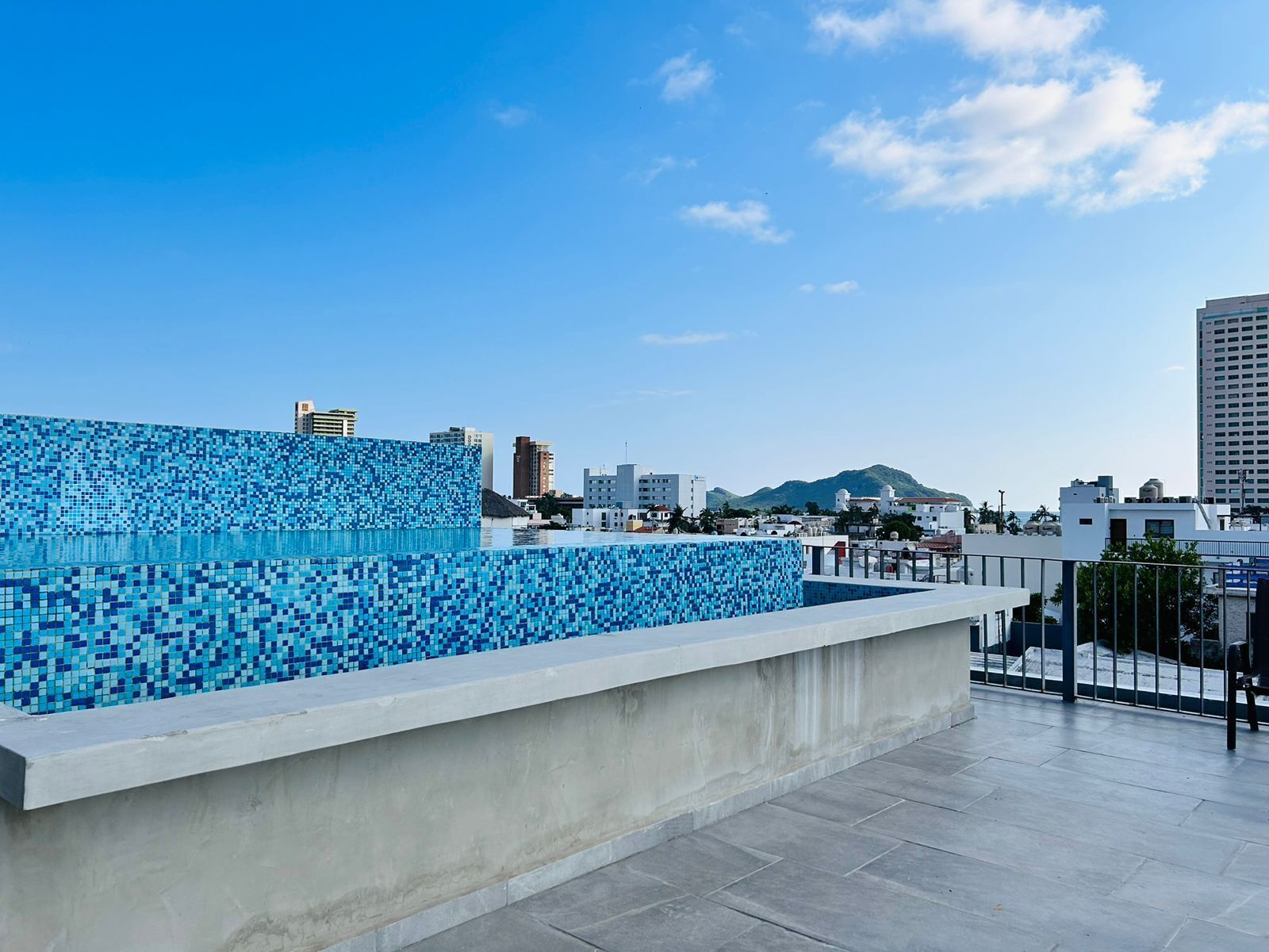 A rooftop pool with a mosaic tile wall, surrounded by a cityscape and mountains in the background.