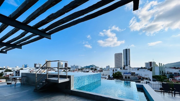 Une piscine sur le toit avec vue sur la ville, un espace salon couvert et une petite table.