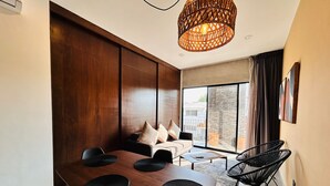 Un salon moderne avec une table à manger en bois, des chaises noires, un canapé avec des coussins, un luminaire en fibres tressées et une grande fenêtre donnant sur l’extérieur.