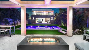 En moderne udendørs terrasse med bålsted, swimmingpool og udsigt til et moderne hus.