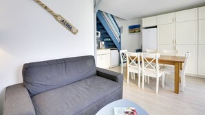 A modern living room with a grey sofa, a wooden wall hanging, a dining area with white chairs and a wooden table, and a kitchen in the background. - Seignosse-océan Villa Patio 4 Personnes (SEIGNOSSE)