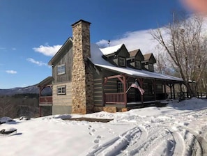 A log cabin with a stone chimney, wooden porch, and a snow-covered landscape.