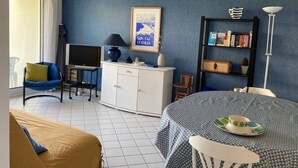 Una stanza con una parete blu, un armadio bianco, una televisione, una libreria, un tavolo da pranzo con una tovaglia fantasia e una finestra con tende.
