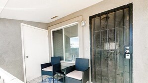 Varanda com duas cadeiras de vime e uma mesa pequena, porta de vidro com grade de metal e tapete estampado.
